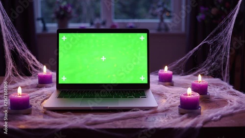 Laptop with Green Screen Display on a Table Decorated for Halloween with Cobwebs and Glowing Purple Candles at Night