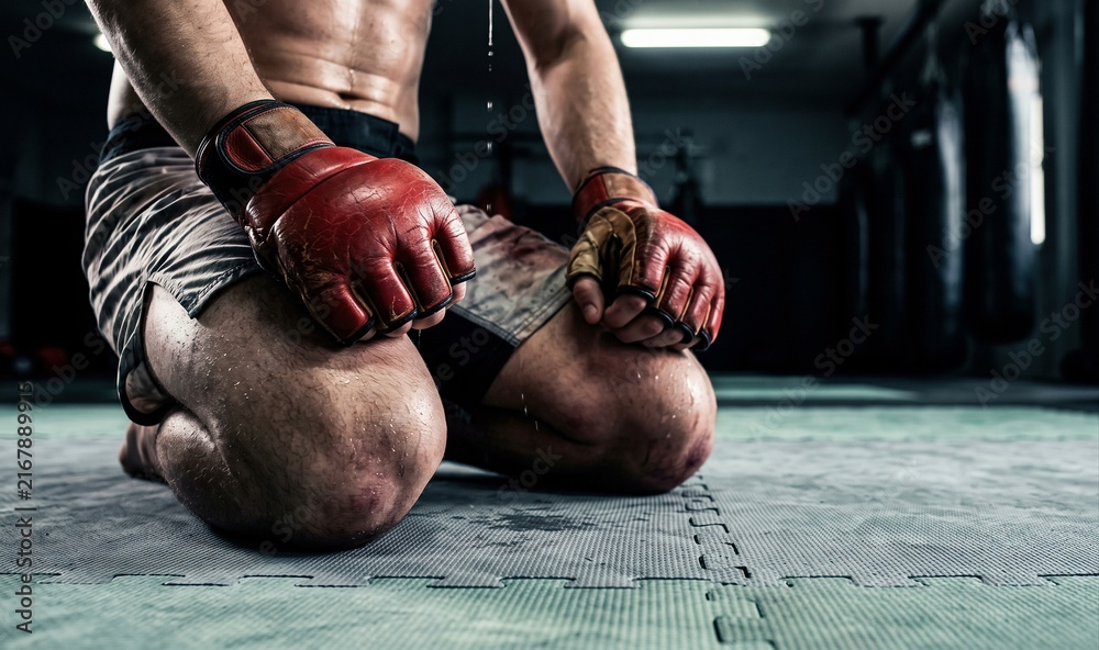 Naklejka premium Fighter kneeling, sweating profusely with open-finger gloves on gym mat, representing intense training and physical exhaustion.