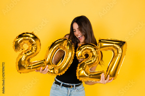 Young woman holding shiny golden 2027 number balloons, smiling broadly with closed eyes and expressing joy against a vibrant yellow background, representing upcoming celebrations