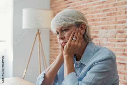 Senior woman experiencing feelings of sadness, loneliness, and depression, sitting indoors with her hands touching her face, contemplating her thoughts in a moment of quiet reflection