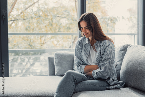 Foto Young woman feeling intense abdominal pain and discomfort, sitting on a sofa at