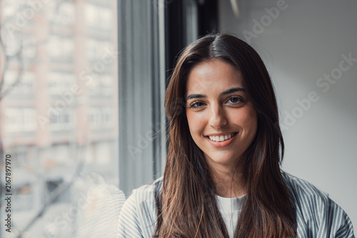 Young woman smiling at the camera, standing indoors by a large window with blurred city buildings in the background, exuding confidence and approachability