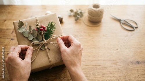 Gift wrapping hands tying twine on brown package with natural decorations on wooden table, for holiday celebration or handmade present.