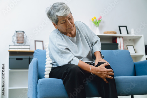 Elderly Asian woman sitting on a sofa at home, holding her painful knee, experiencing joint discomfort and mobility problems.