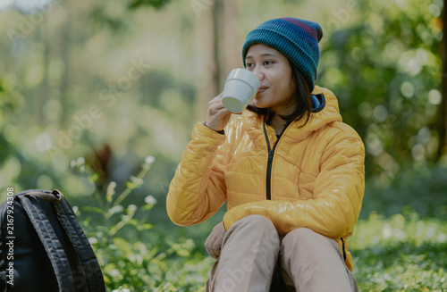 Attractive young active Asian female in a yellow puffer goose down jacket drinking a coffee cup sits on the camp ground area in the nature outdoor park.