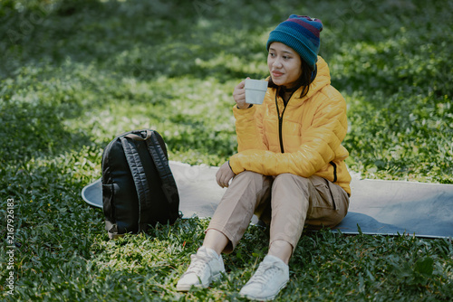 Attractive young active Asian female in a yellow puffer goose down jacket drinking a coffee cup sits on the camp ground area in the nature outdoor park.