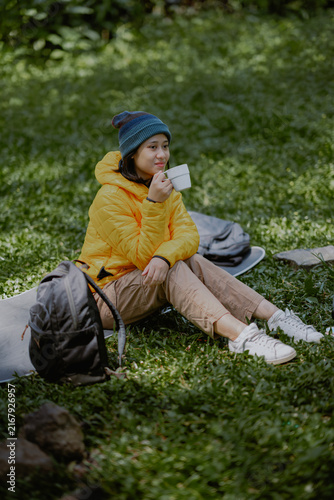 Attractive young active Asian female in a yellow puffer goose down jacket drinking a coffee cup sits on the camp ground area in the nature outdoor park.