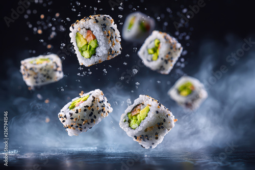 Several sesame-coated uramaki rolls with avocado filling suspended in mid-air above a wet reflective surface, surrounded by water droplets and drifting mist.
