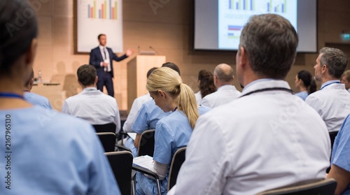 Medical audience of healthcare professionals in white coats and scrubs attending seminar with speaker presenting charts on screen in modern conference auditorium setting
