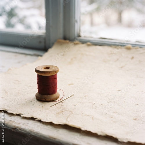 Vintage wooden spool with red thread and sewing needle on handmade textured paper by window, cozy handicraft craft concept.