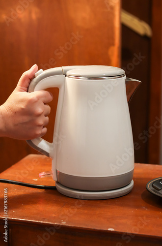 Electric kettle on a wooden table, held by a woman's hand, used for boiling water in a kitchen, office pantry, or hotel room.