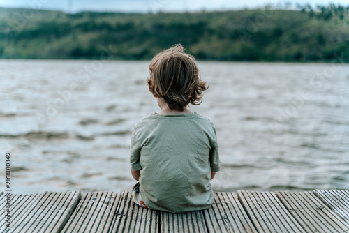 Lonely child sitting on a wooden dock looking out at a calm lake