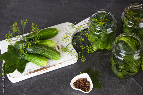 Cuadro en lienzo Cucumbers in glass jars and on a cutting board, dill, garlic, and spices on a white wooden background