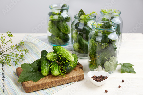 Cucumbers and herbs in jars on a table with cutting board and spices for pickling