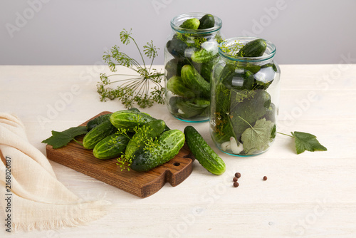 Cucumbers and herbs sit on a cutting board beside jars filled with additional cucumber and leaves.