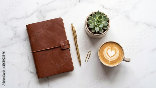 Brown leather journal placed on white marble surface alongside golden pen succulent plant and ceramic cup of hot coffee