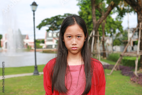 A young Asian girl with long dark hair, She is in an outdoor park setting with green grass, trees, and a blurred background featuring a fountain.