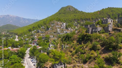 Kayakoy, Turkey. Hundreds of abandoned stone house ruins cascading down a forested mountainside, aerial drone view under clear blue sky with distant peaks. Aerial View