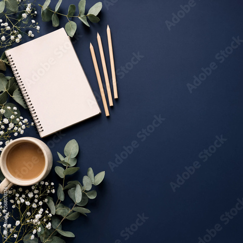 Flat lay of open notebook with pencils, eucalyptus leaves, and coffee on dark blue background