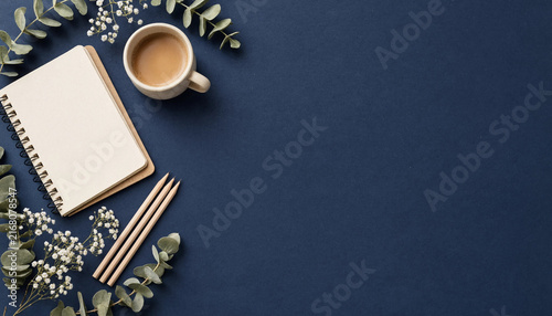 Top view of a notebook, coffee cup, pencils, and eucalyptus leaves on a dark blue background