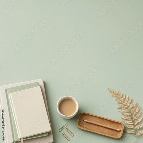 Minimalist flat lay with notebooks, coffee cup, and fern on sage green background