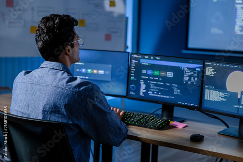 Plakat w ramie Machine learning engineer reviewing AI model dashboard and system performance at computer workstation while analyzing technical metrics, software status, data visualization and model results.