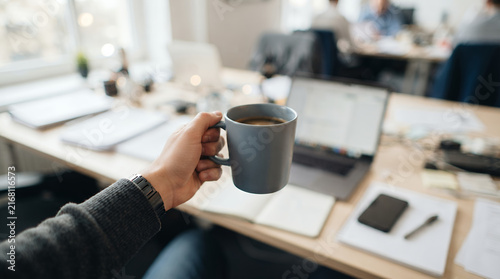 First-person perspective of hand holding coffee mug in office, busy corporate workspace with laptop and documents, morning energy concept