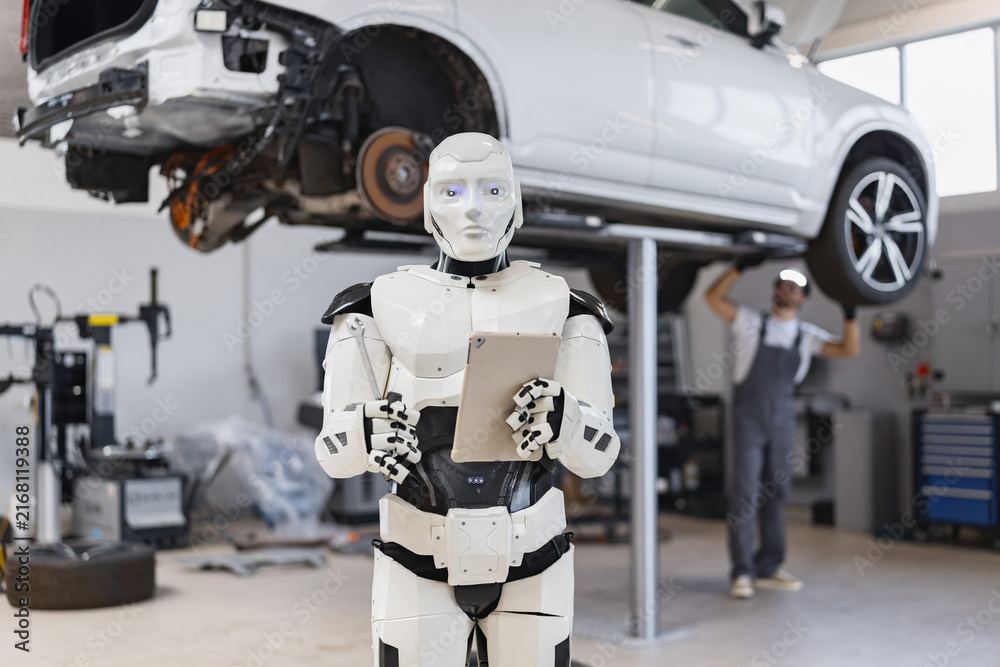 Fototapeta premium A humanoid robot with glowing eyes holds a tablet and a wrench while a mechanic works on a car lifted on a hoist in a garage