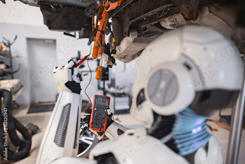 A humanoid robot uses a multimeter to test electrical wiring on a vehicle chassis in a workshop setting