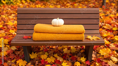 Autumn park bench with cozy knit blanket and pumpkin on fallen leaves in golden sunlight
