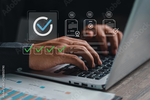 Close up of hands typing on laptop with ISO standard certification documents and green checkmark checklist icons