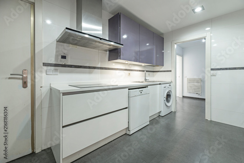 Modern white kitchen with lavender cabinets, stainless hood, dishwasher, and washing machine leading to a tiled hallway.