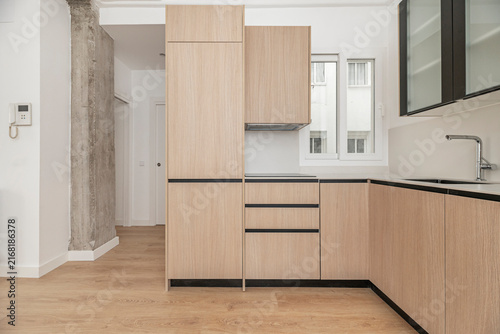 L-shaped oak kitchen with concrete pillar, intercom panel, corner sink, and window flanked by tall wooden cabinetry.