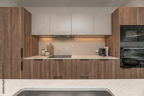 Symmetrical walnut kitchen with warm under-cabinet lighting, induction cooktop, coffee machine, and glimpse of dining chair beyond.