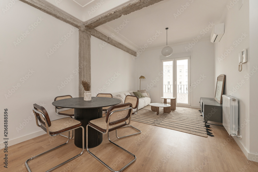 Fototapeta premium Living-dining room with exposed concrete beam, black round table, cane chairs, sofa, and balcony view