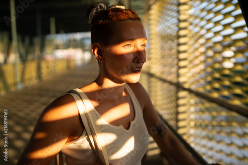 A white woman stands in sunlight and shadow on an urban bridge