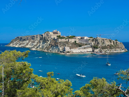 Panoramic View of San Nicola Island Fortress and Abbey Framed by Pine Trees from San Domino, Tremiti Archipelago, Italy – Turquoise Sea with Boats Under Sunny Blue Sky