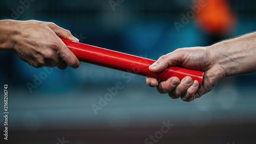 Athletes passing a red relay baton during a track race. This imagery represents teamwork, professional success, and seamless business transition concepts.