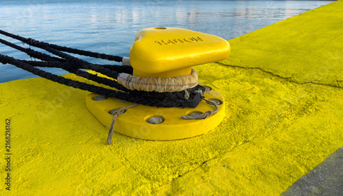 Obraz na płótnie Black mooring ropes around yellow bollard on Chios harbor quay beside calm water