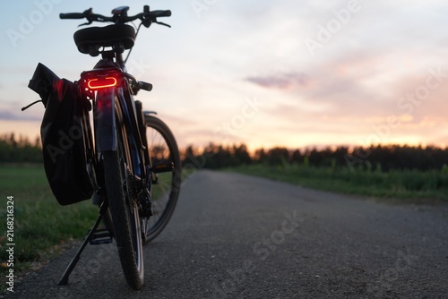 Bicycle with illuminated rear light and pannier bag parked on a bike path at sunset, with open countryside, warm evening sky and generous copy space on the right