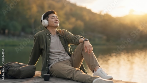 Young man relaxing with headphones by lake listening to music at sunset