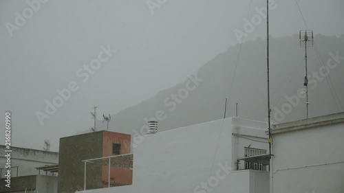 Heavy rain and strong precipitation falling against background of low rise European Spanish buildings and high mountain in distance. Powerful rains during cyclone passage causing floods, high water.