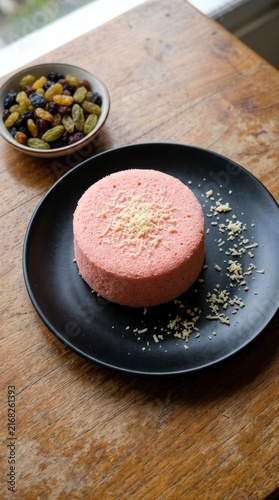 Pink steamed cake topped with grated cheese on a black plate, bowl of mixed raisins on wooden table.