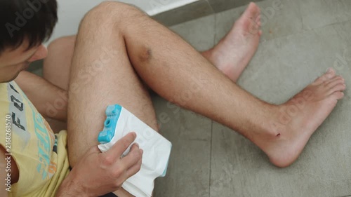 Man applying cold pack wrapped in napkin to thigh hematoma at home. Self care cooling of injured spot, first aid at home after trauma, top view. Improvised pain relief for bruise after impact.