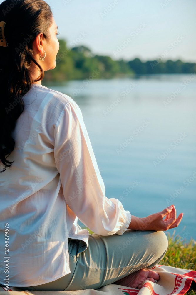 Naklejka premium Vertical image of young adult Asian woman practicing yoga and meditating by river. Scene represents mental wellbeing and mindfulness, perfect for wellness and health campaign