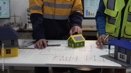 A close-up shot of an architect and a civil engineer discussing blueprints and a house model in an office at night.