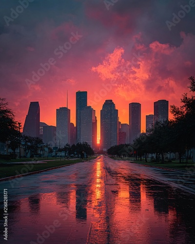 A city skyline silhouetted against a dramatic, colorful sunset reflected in a wet street after rainfall. Trees line the road