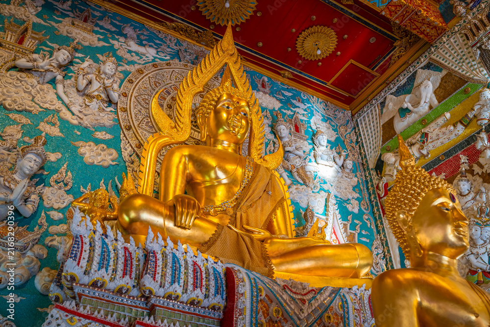 Fototapeta premium Golden Buddha statue inside the hall of Wat Sri Uthumphon in Mueang Nakhon Sawan District, Nakhon Sawan, Thailand, surrounded by ornate white relief wall art and celestial angels.