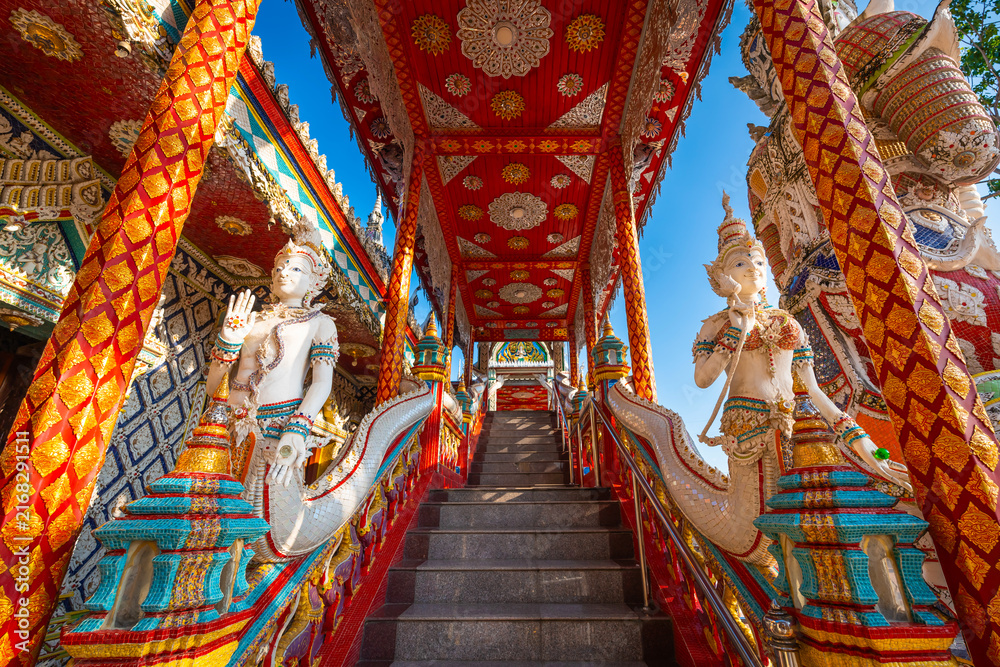 Fototapeta premium Ornate pavilion staircase flanked by white angel guardian statues at Wat Sri Uthumphon in Mueang Nakhon Sawan District, Nakhon Sawan, Thailand, featuring rich red and gold traditional Thai art.