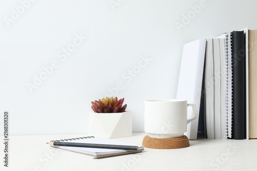 Fotografía Stylish workspace with wooden desk, office supplies and white cup near white wall